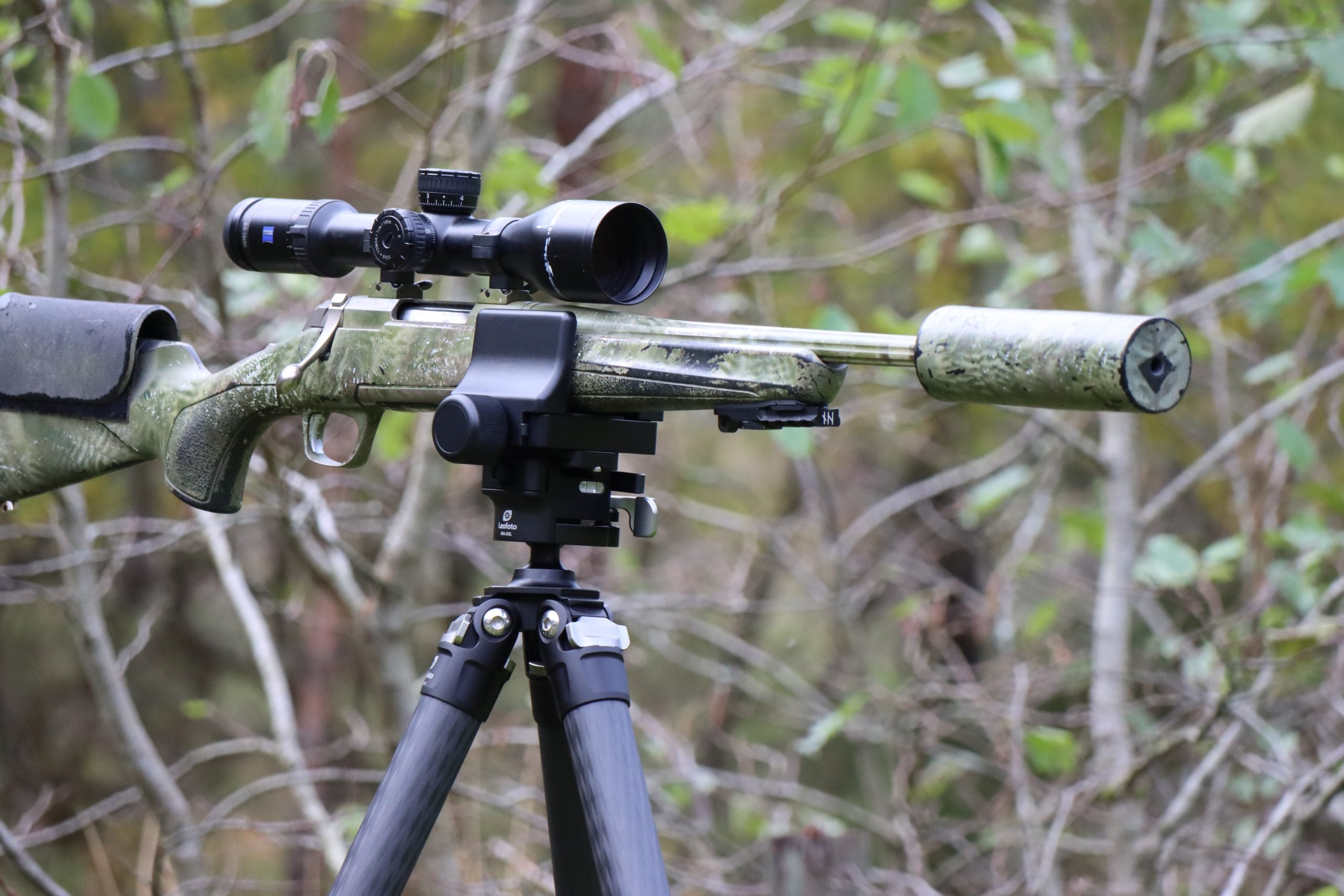 Leofoto GS-3 Equipment Support Mount securing a scoped rifle on a tripod in a forest setting.