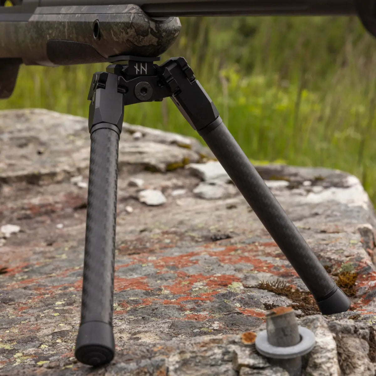Close-up of a NorneHunt GEN II bipod mounted beneath a camouflage rifle stock, with its feet resting on rock.