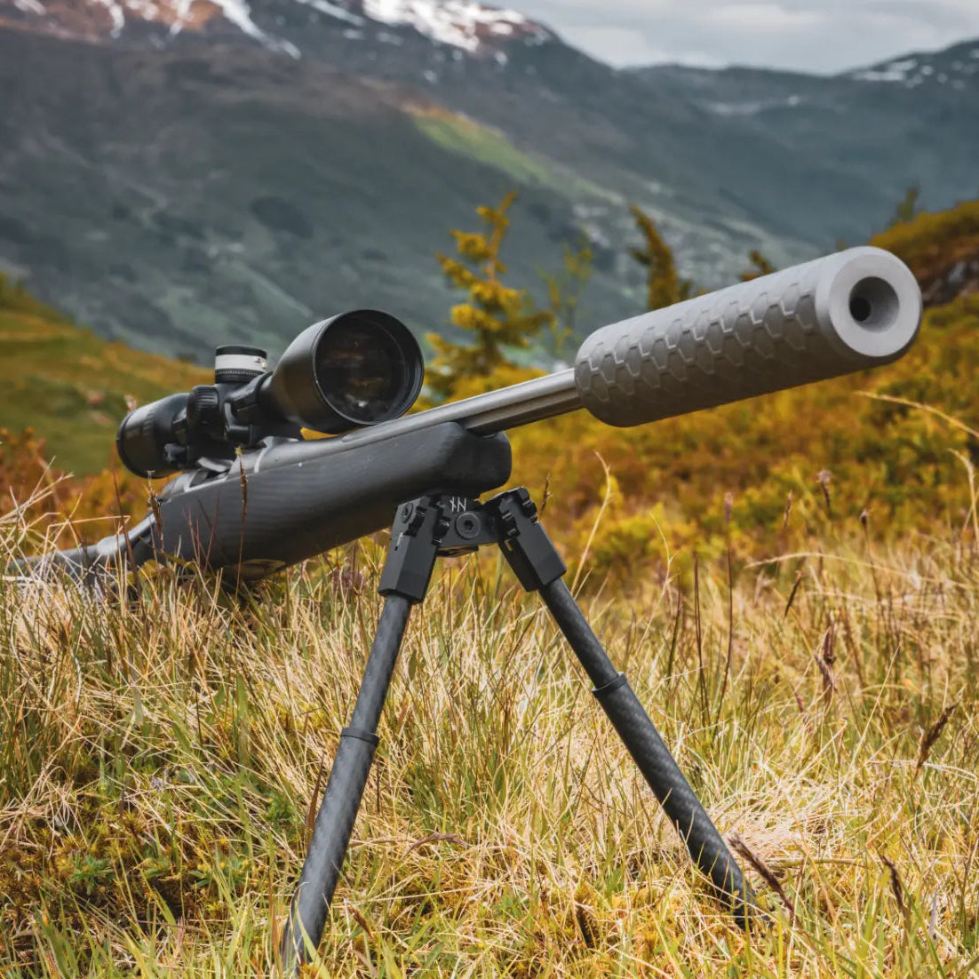 Scoped hunting rifle supported by a NorneHunt GEN II bipod on a grassy hillside with mountains behind.