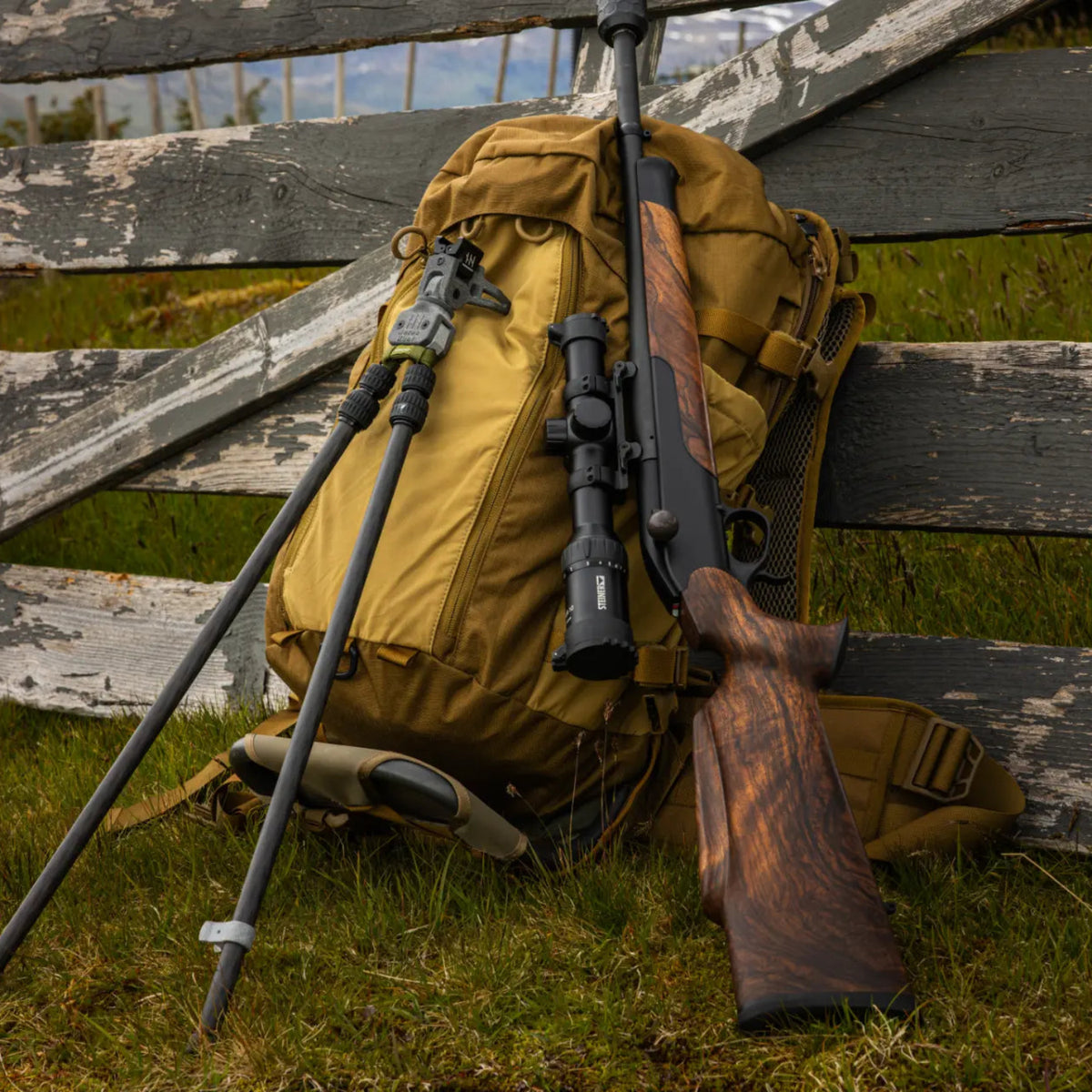 NorneHunt shooting sticks beside a scoped wooden-stock rifle and yellow rucksack against a wooden bench.