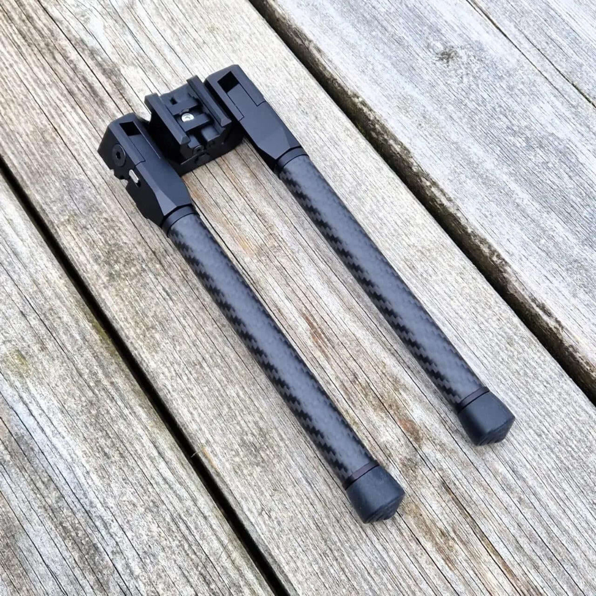 Folded NorneHunt SvartR bipod on wooden boards, showing its carbon fibre legs and Picatinny clamp.